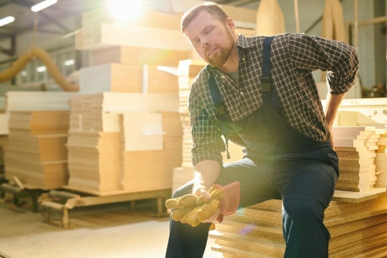 Manual worker with closed eyes sitting on stack of wooden plank and massaging lower back while feeling pain in back possible Virginia workers compensation claim.