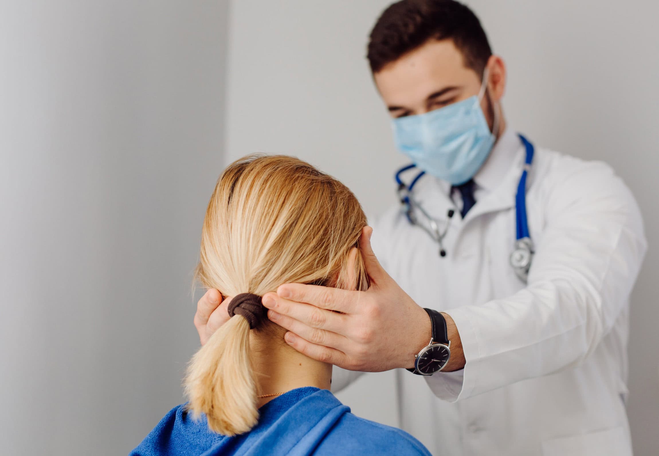 A doctor wearing a face mask examines a patient’s neck and head during a medical checkup.