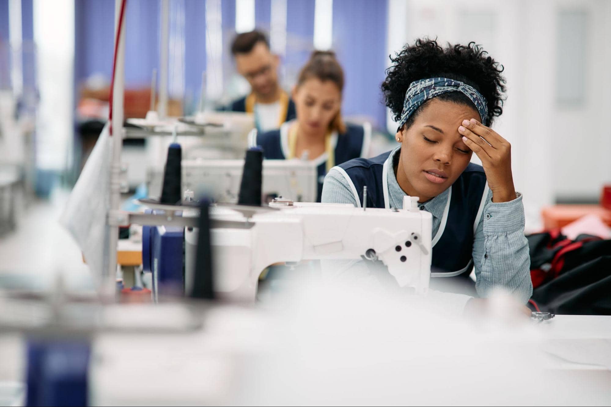 A tired worker sitting at a sewing machine in a factory, resting her head on her hand, with other workers focused in the background.