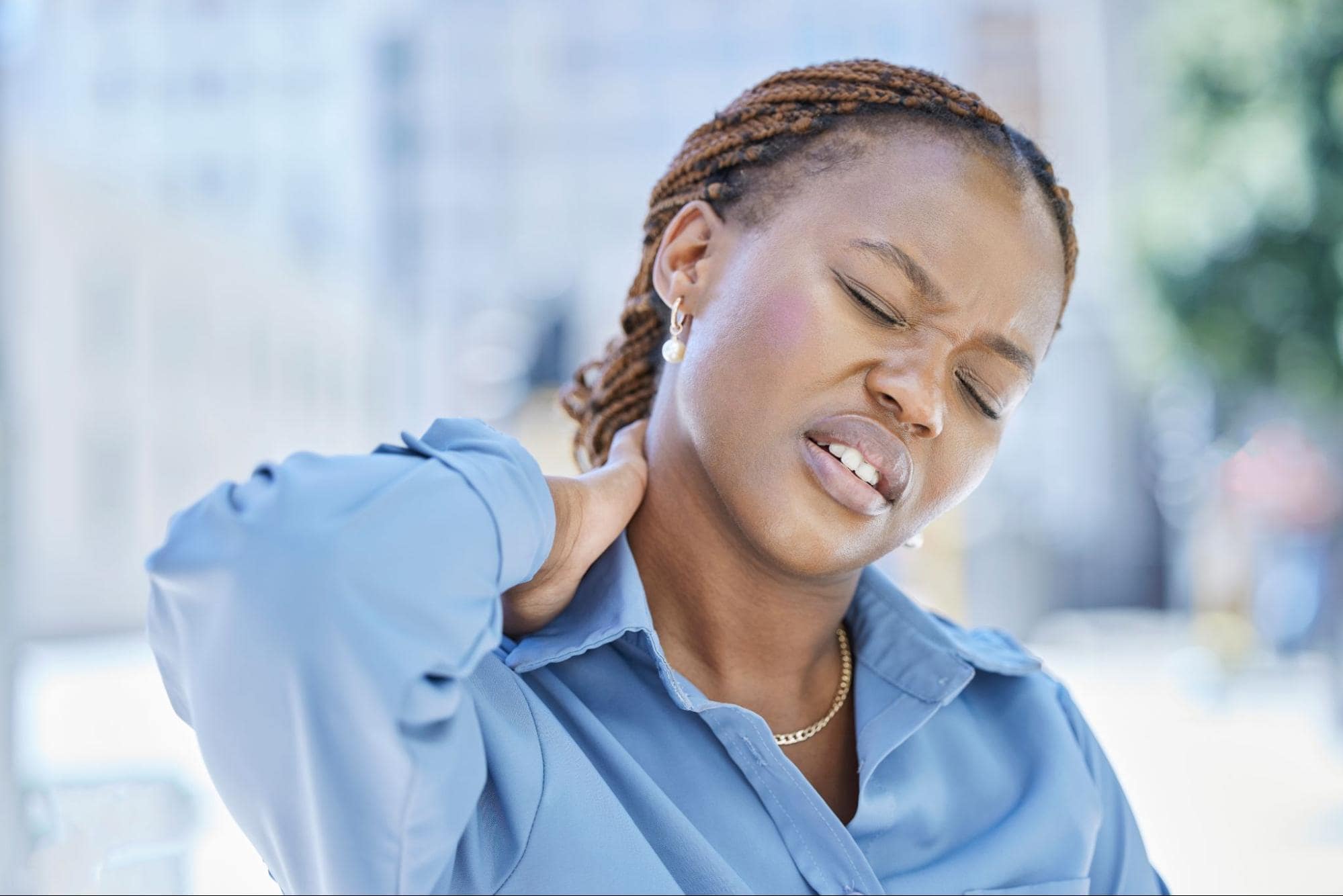 A woman in a blue blouse grimaces and holds the back of her neck with one hand, appearing to be in pain or discomfort.