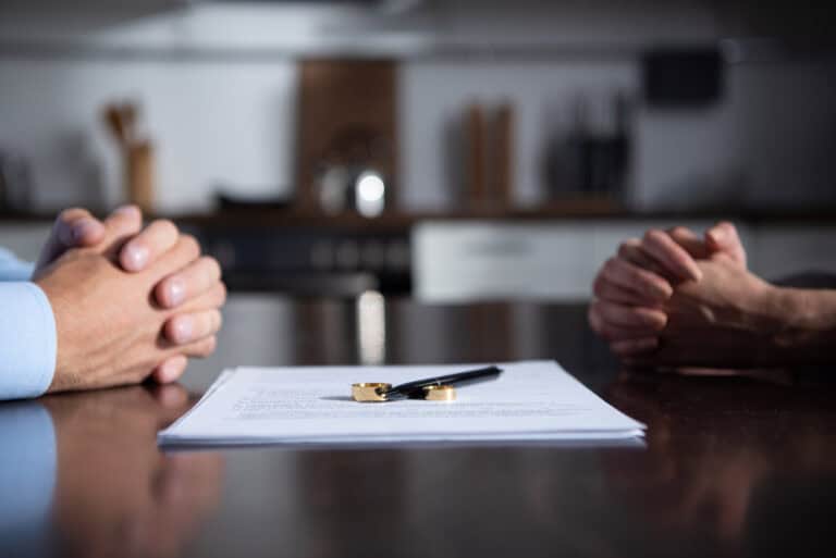 Close-up of two people sitting across from each other at a table with hands clasped, a document between them, and two wedding rings placed next to a pen, symbolizing divorce or separation.