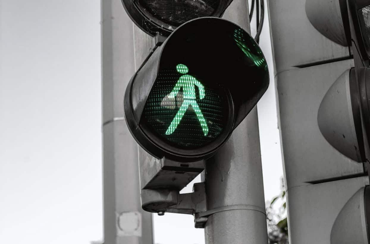 Green pedestrian walk signal illuminated on a crosswalk traffic light mounted to a metal pole at an urban intersection.