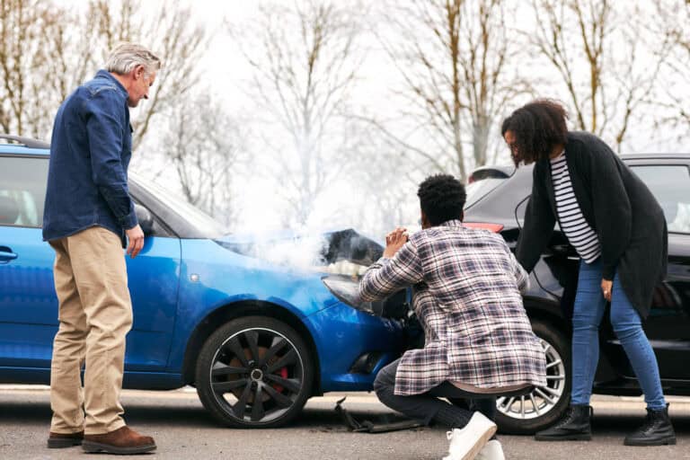 Three people inspect vehicle damage after a car accident, one person photographing the smoking front end of a blue car involved in a collision.