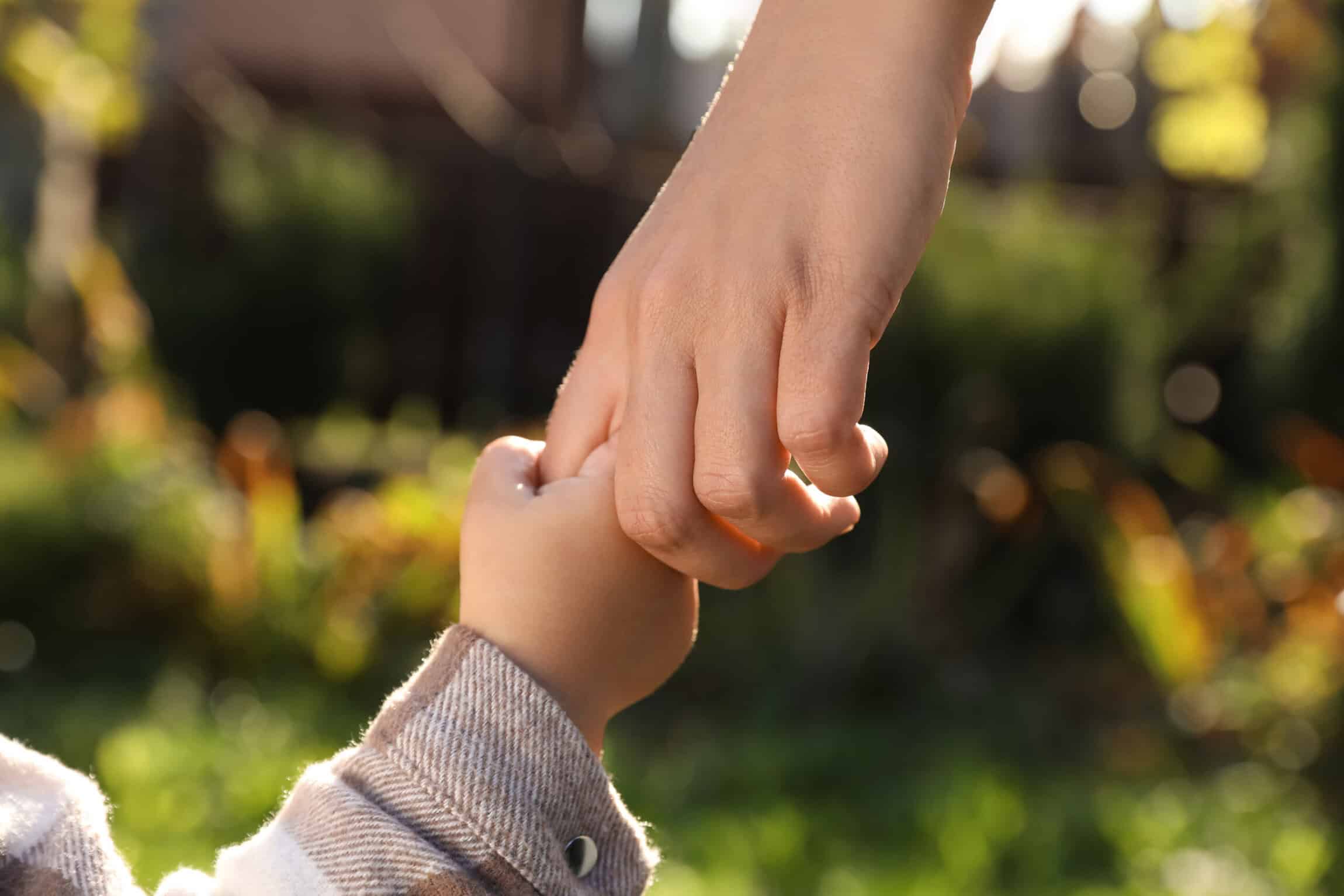 Parent holding a young child’s hand outdoors in warm sunlight, symbolizing care, support, and family connection